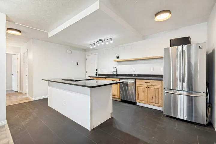Open-concept kitchen featuring dark tile flooring, a center island with a dark countertop, light wood cabinetry, stainless steel appliances, and a floating wood shelf