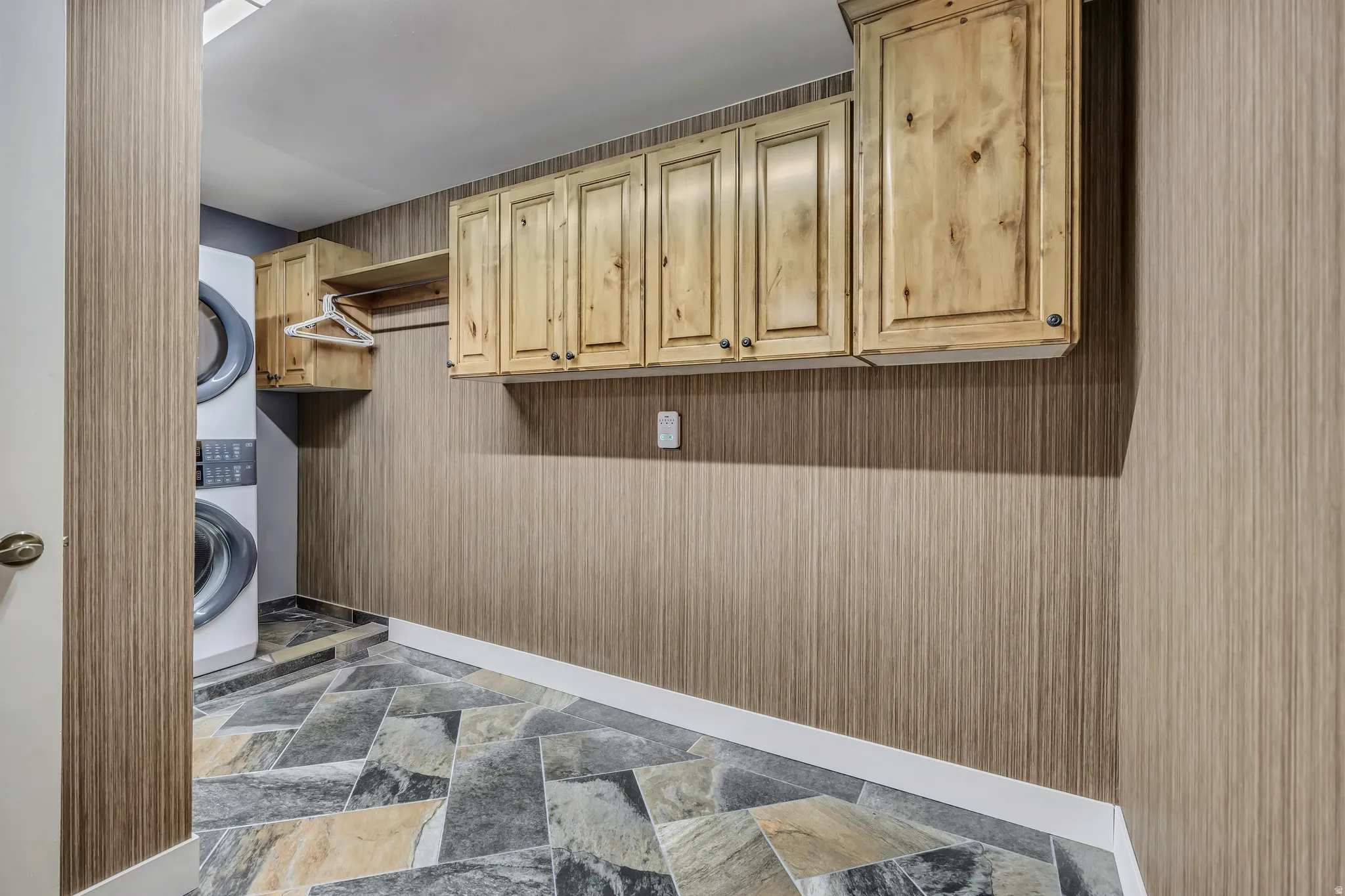 Laundry area featuring custom wood cabinetry, a built-in hanging rod, stacked front-load appliances, variegated stone-finish flooring, and wood-grain wall paneling