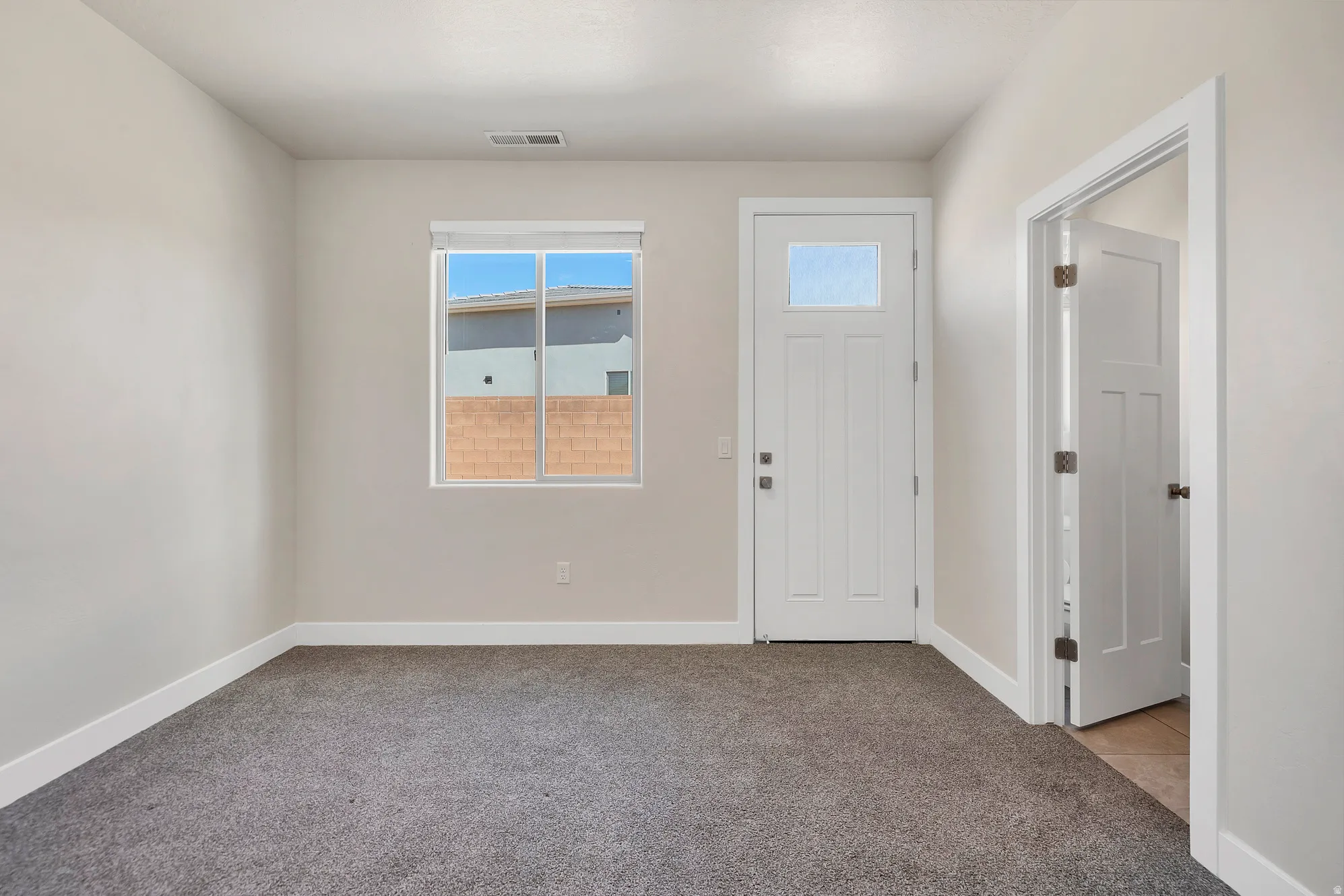 Neutral-toned interior room featuring a white paneled entry door with frosted glass, a large window, gray carpet flooring, and white baseboards