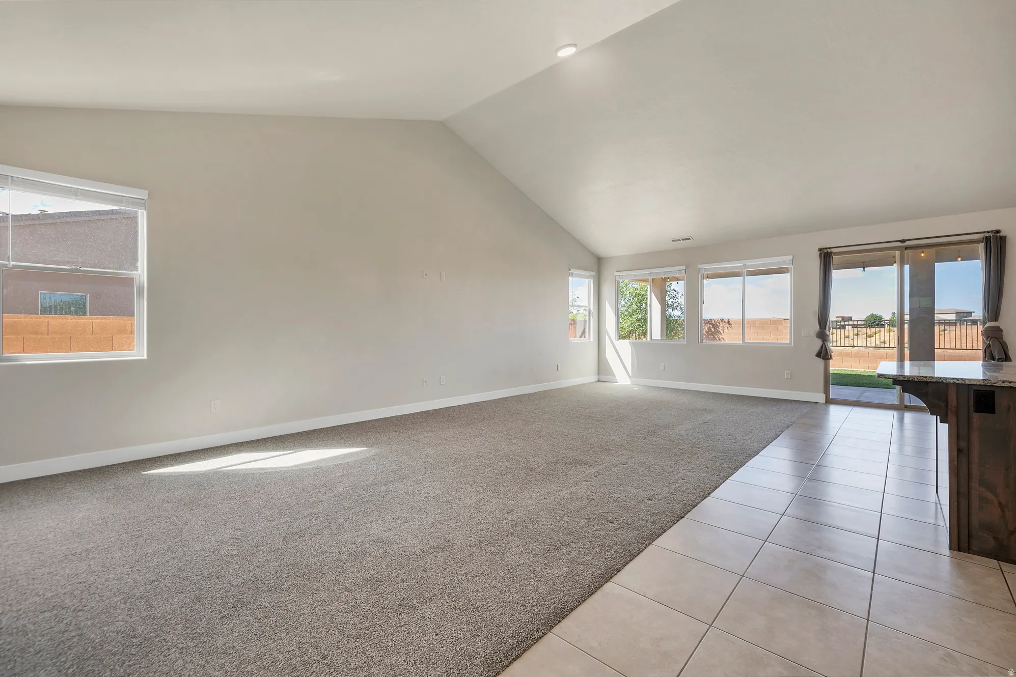 Spacious open-concept room featuring vaulted ceilings, multiple windows, plush carpeting, and a sliding glass door providing exterior access