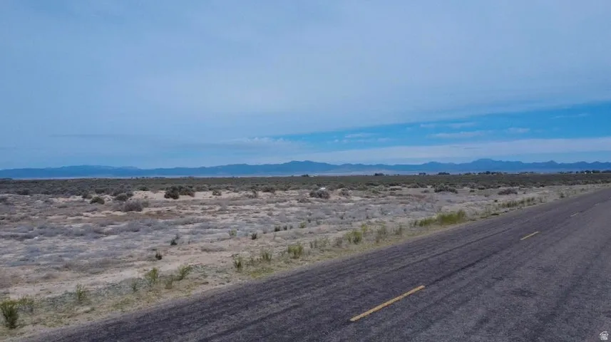 Expansive rural land featuring a paved road with yellow markings, distant mountain ranges, and scattered desert vegetation under a wide sky