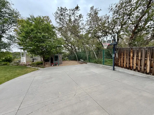 Spacious concrete patio featuring a basketball hoop, surrounded by mature trees and a wooden privacy fence