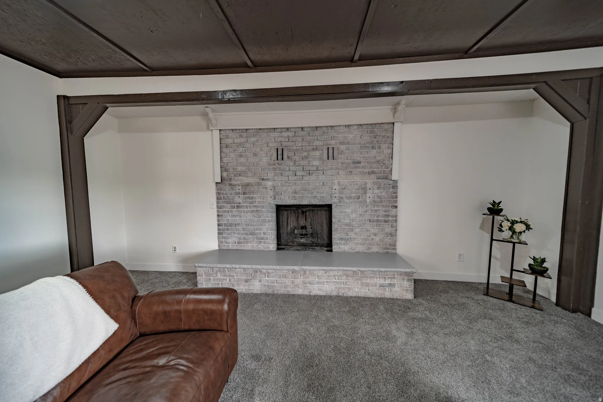 Living area featuring a painted brick fireplace with a raised hearth, wood-beamed ceiling, and wall-to-wall carpeting