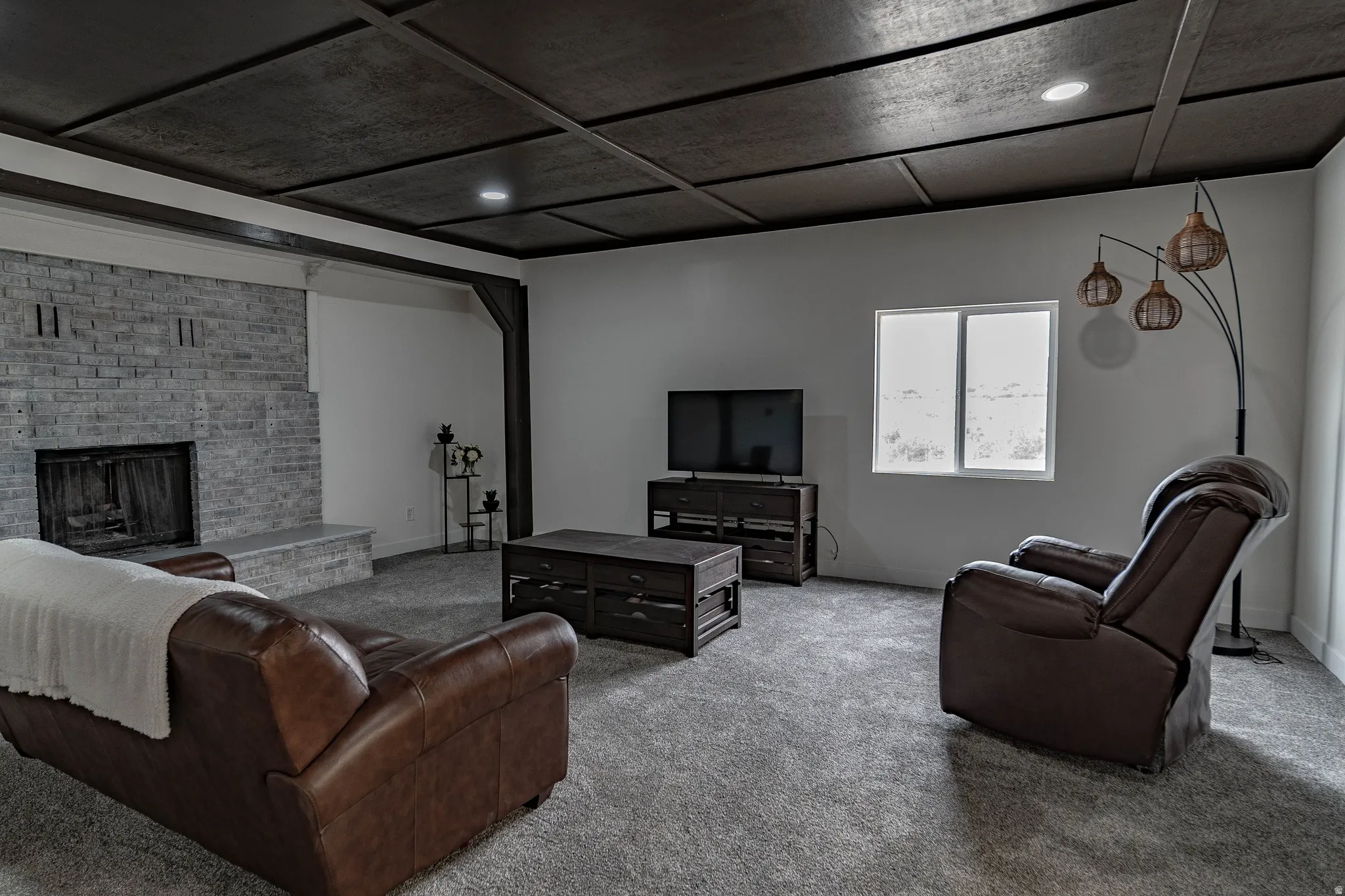 Tray ceiling with recessed lighting and dark wood-finish details, light gray brick fireplace with stone hearth, light gray wall finishes, and gray carpet flooring