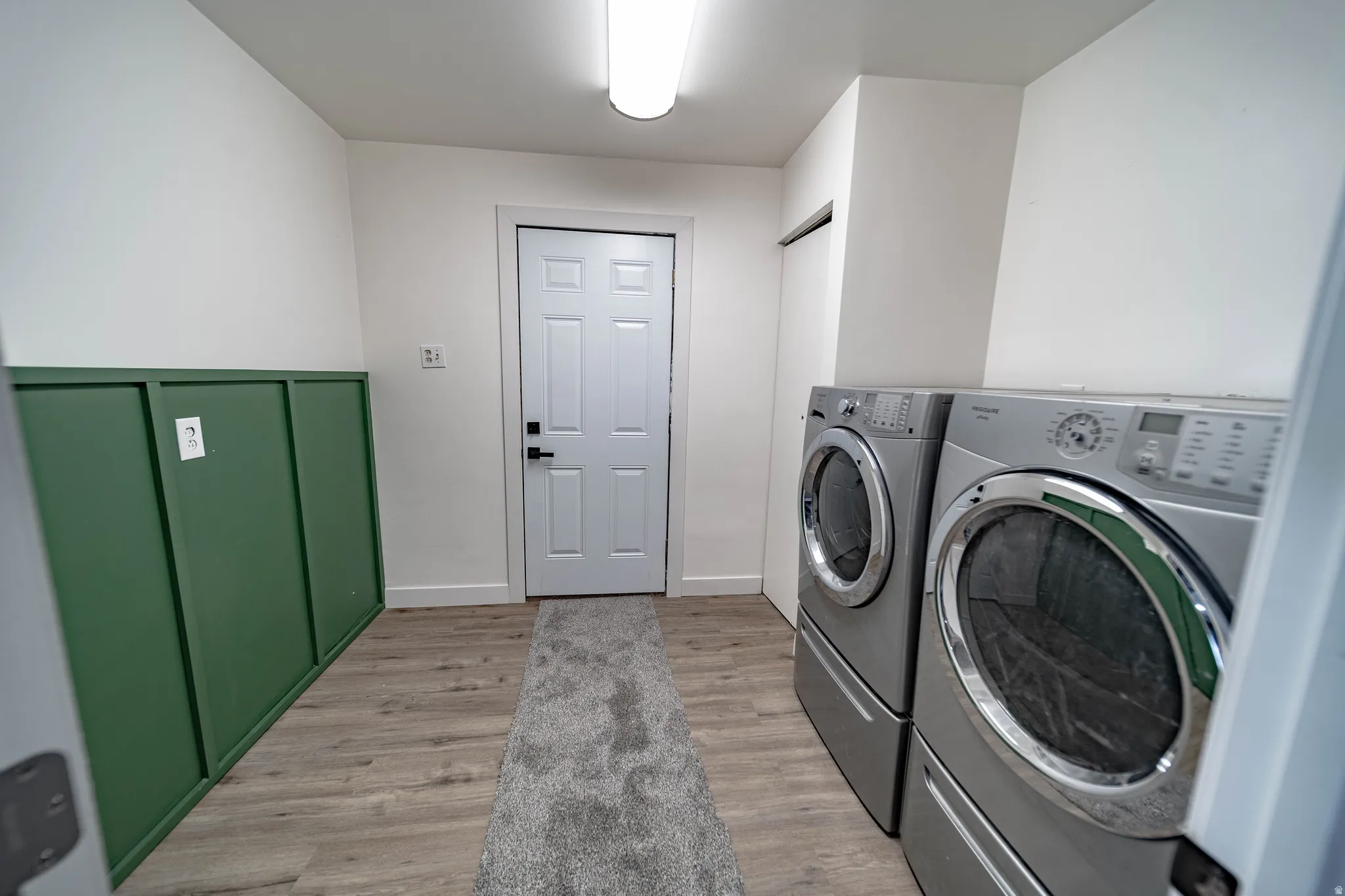Laundry area featuring wood-finish flooring, white walls with green wainscoting, a white paneled door, and a flush-mount ceiling light