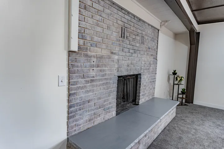 Exposed brick fireplace with a hearth, white painted walls, and textured carpeting