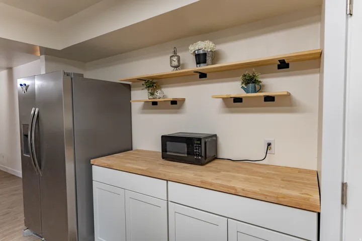 Stainless steel side-by-side refrigerator, butcher block countertop, white shaker-style cabinetry, and natural wood floating shelves