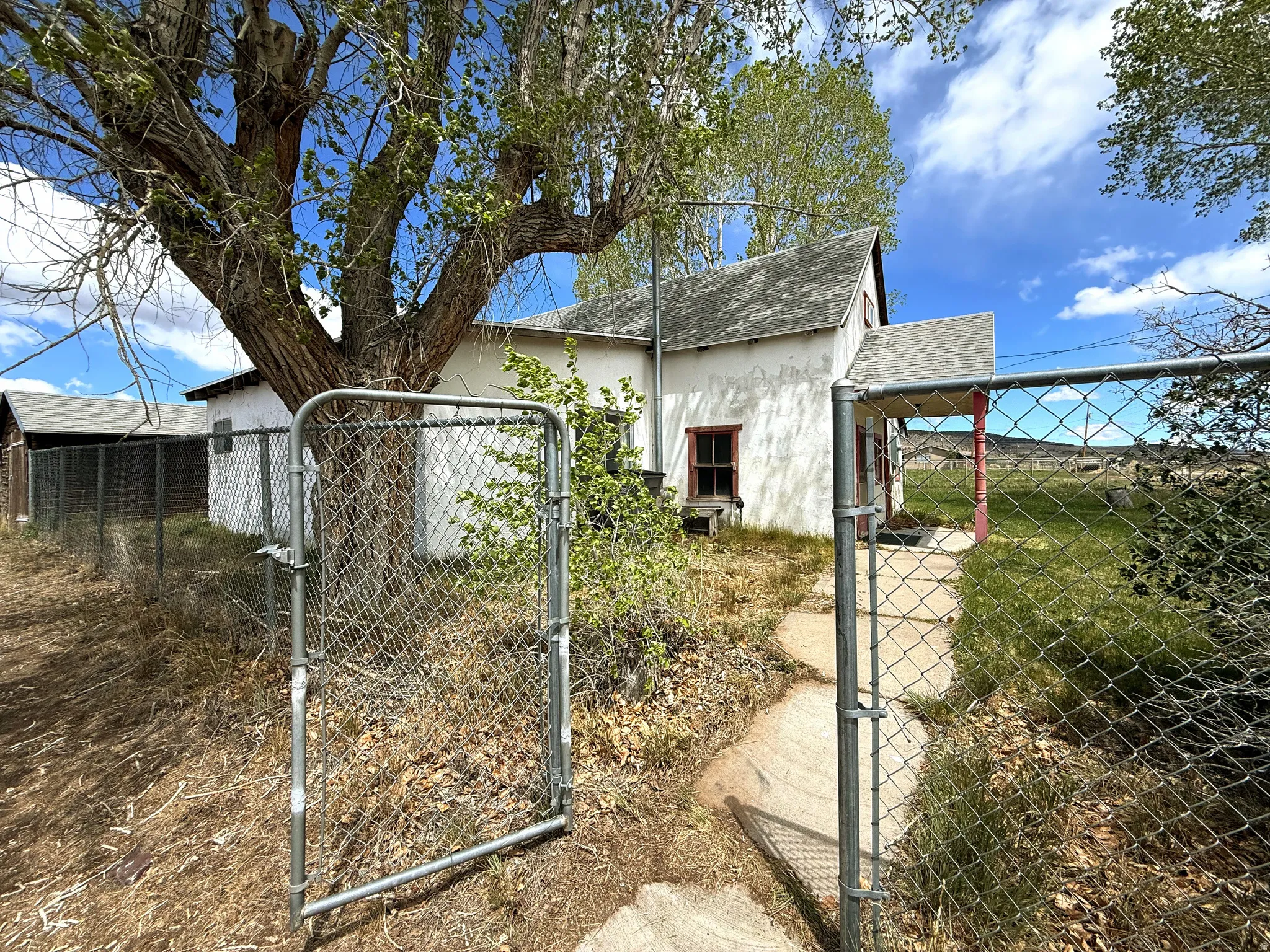 Chain-link fence with an open gate, bordering a gravel path leading to a white stucco dwelling