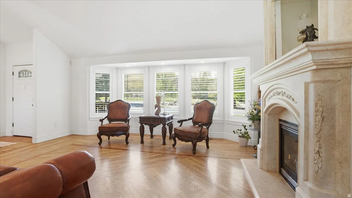Expansive living area featuring light wood-finish flooring, a decorative fireplace with ornate mantel, and a multi-panel bay window with plantation shutters