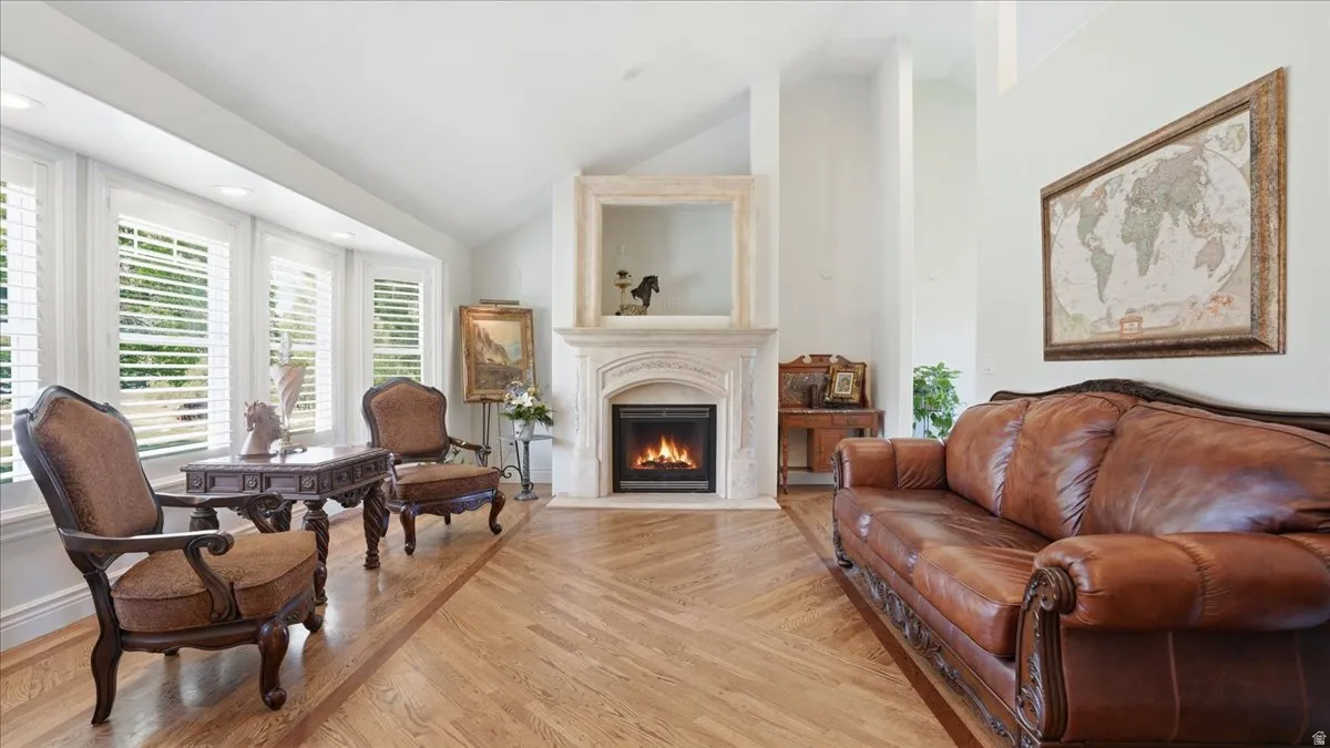 Living area featuring a focal fireplace with an ornate mantel, a bay window with plantation shutters, vaulted ceilings, and wood-finish flooring with a decorative inlay