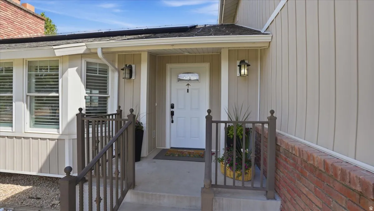 Front entry featuring a white paneled door, two exterior sconces, board and batten siding, brick detailing, and a metal railing