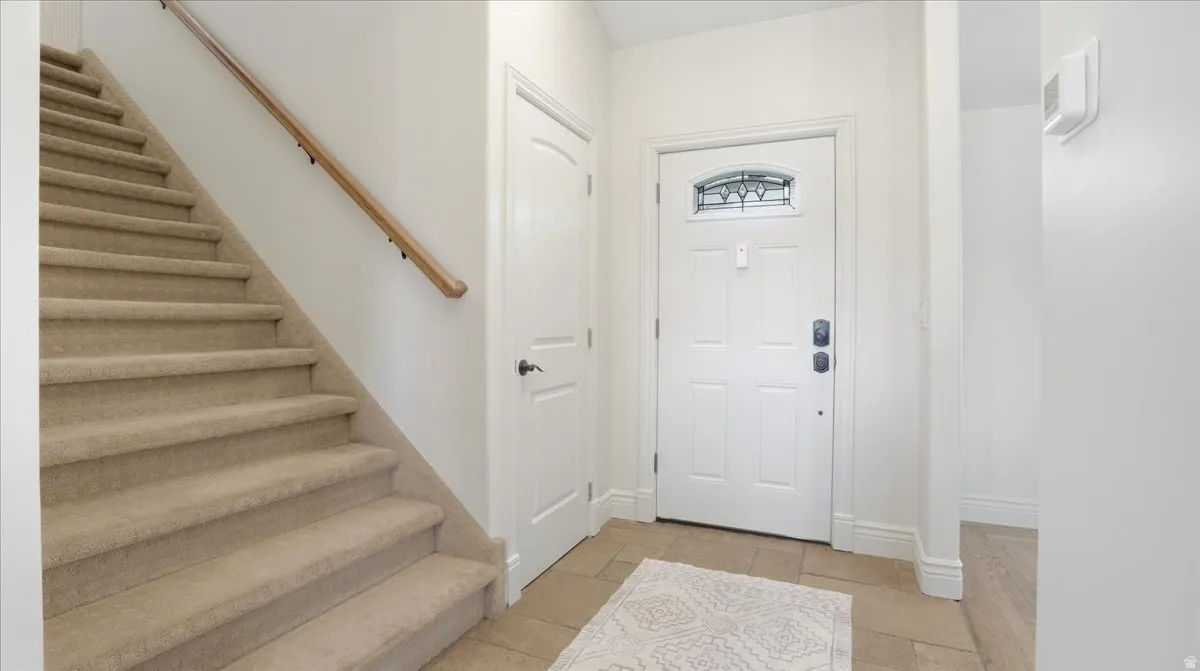 Entry foyer featuring a paneled door with decorative glass, beige tile flooring, and a carpeted staircase with a wood handrail