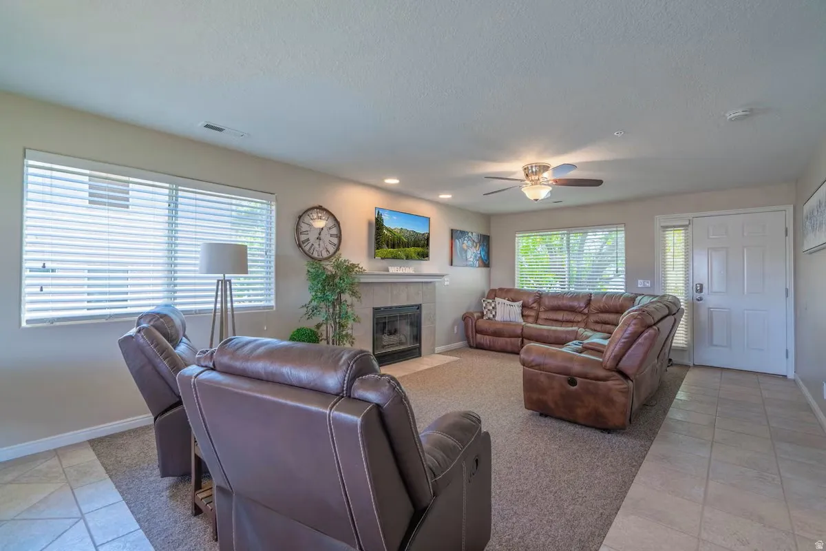 Living room featuring a fireplace with a stone surround and mantel, large windows with blinds, a ceiling fan with integrated lighting, and mixed flooring with carpet and tile