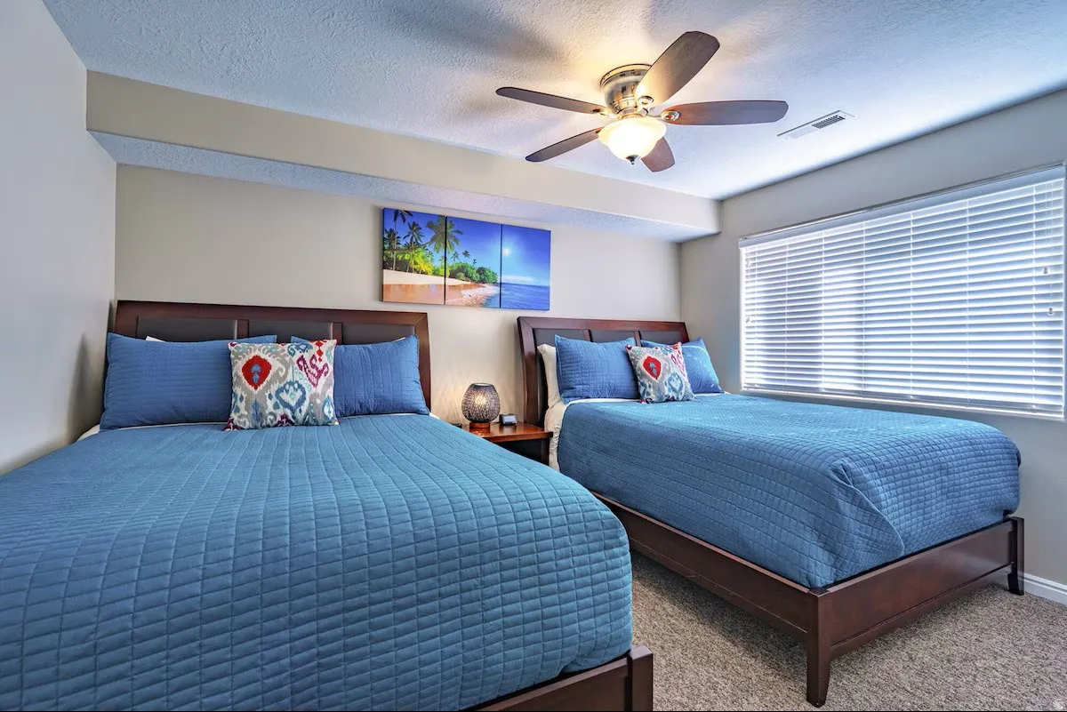 Bedroom featuring two wood-frame beds, textured carpeting, a ceiling fan with integrated lighting, and a window with horizontal blinds