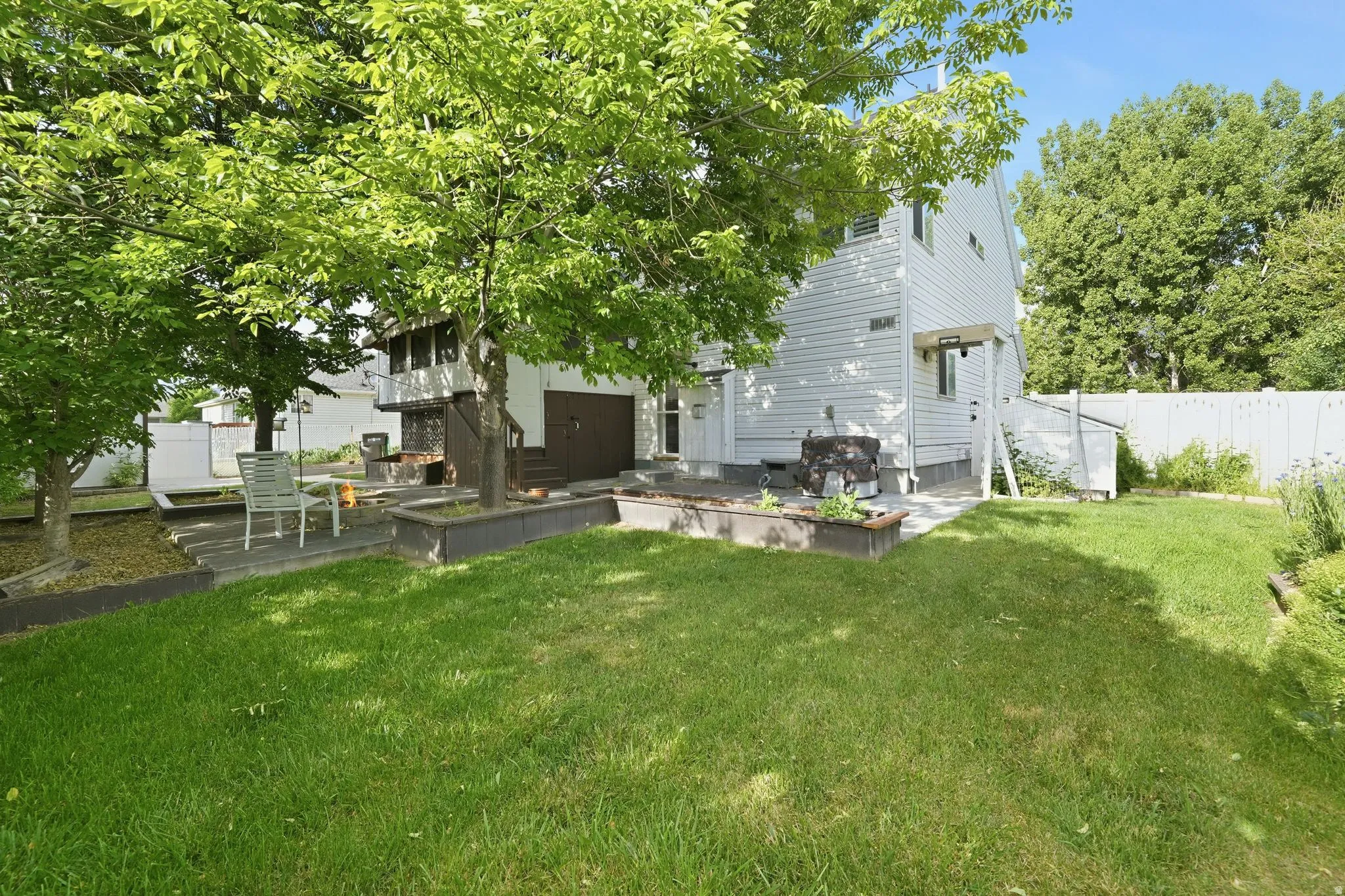 Expansive rear yard featuring mature trees, a manicured lawn, a white privacy fence, a concrete patio, and dark-stained wood planter boxes