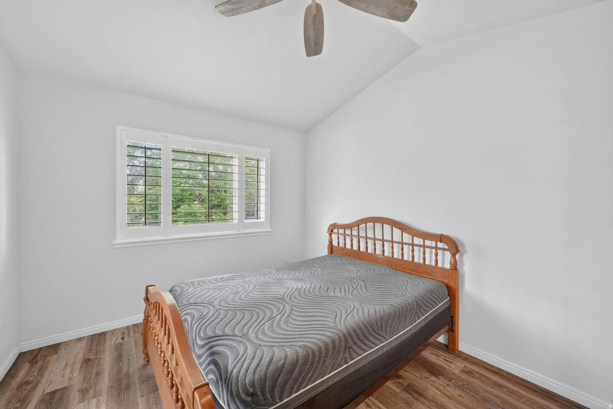 Vaulted ceiling bedroom featuring wood-finish flooring, a three-panel window with plantation shutters, and a ceiling fan