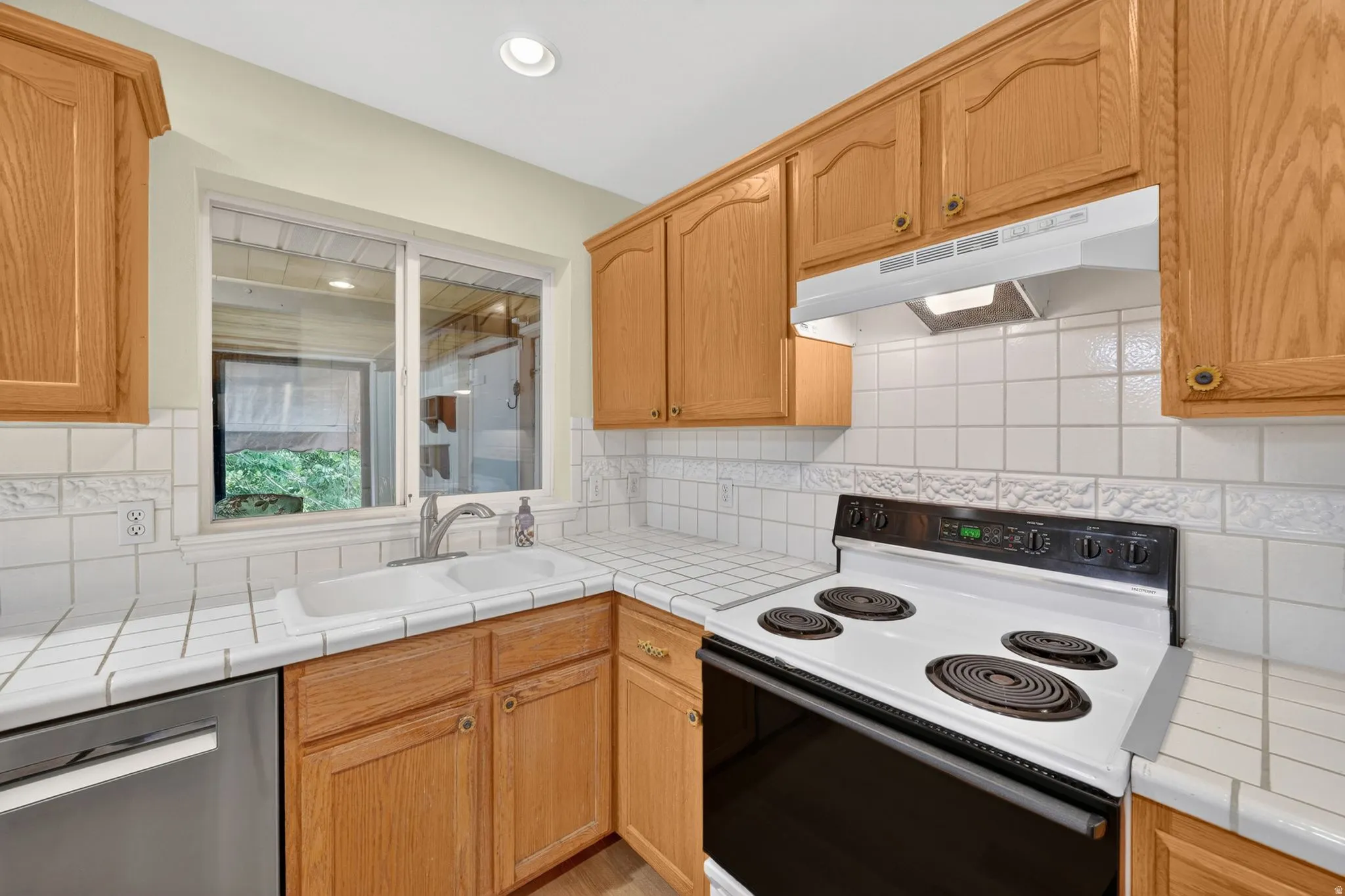 Kitchen featuring wood cabinetry, tile countertops, tile backsplash, stainless-steel sink, and a white electric range with a range hood