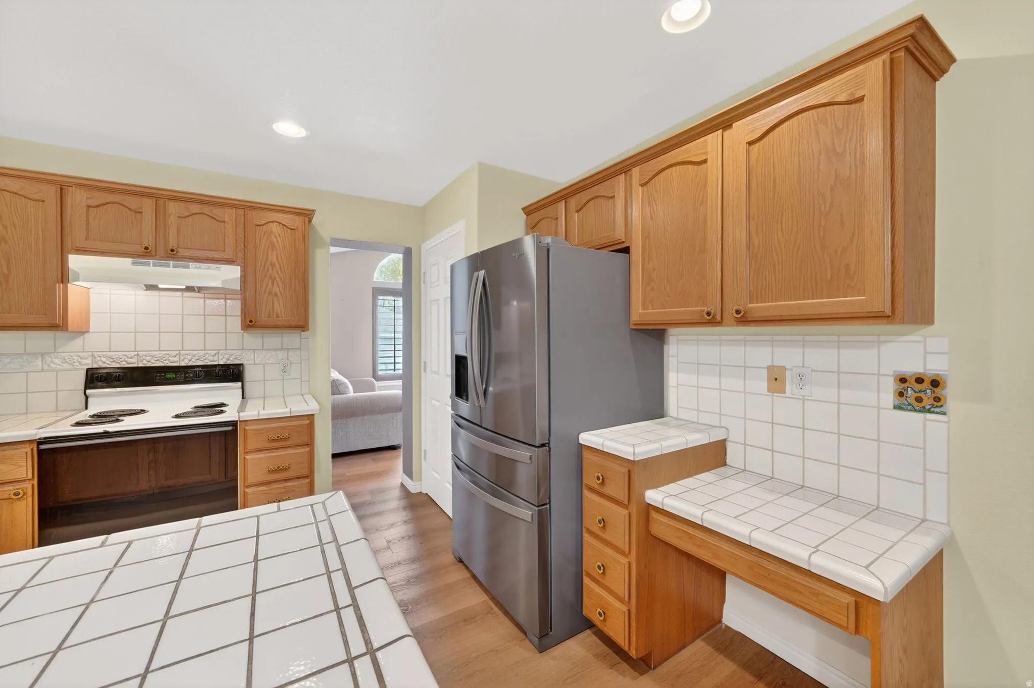 Kitchen featuring wood cabinetry, white tile countertops and backsplash, stainless steel refrigerator, and built-in desk area