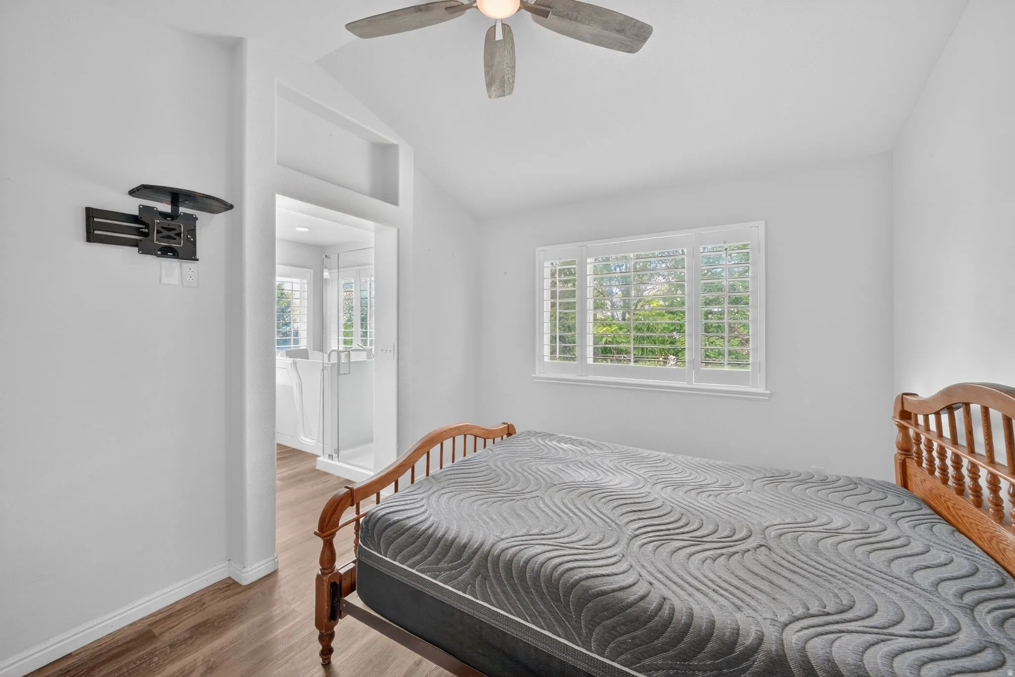 Bedroom featuring wood-finish flooring, a ceiling fan, a window with white shutters, and a built-in television mount