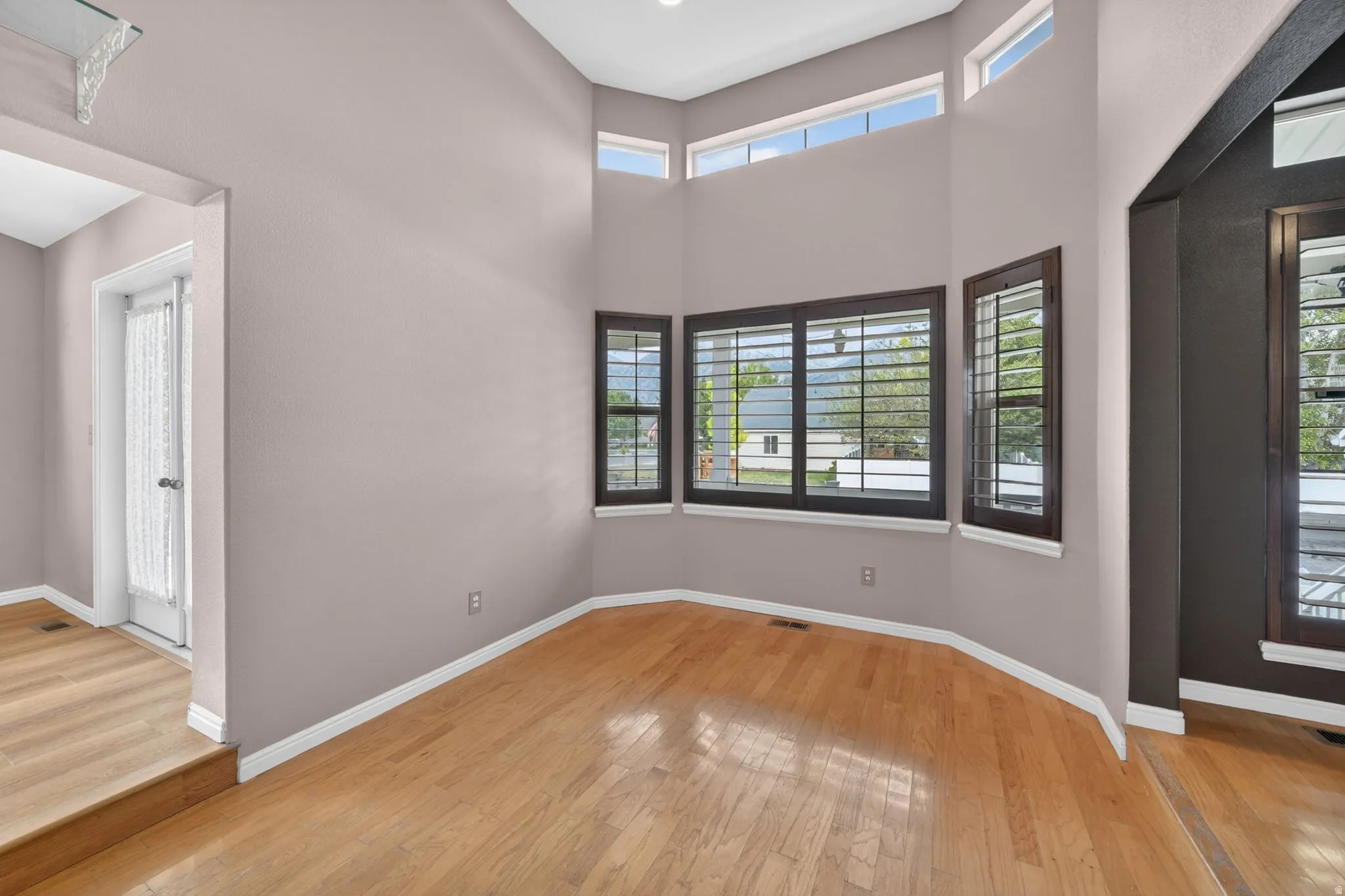 Interior space featuring light wood-finish flooring, high ceilings, and a bay window configuration with dark wood trim and horizontal blinds
