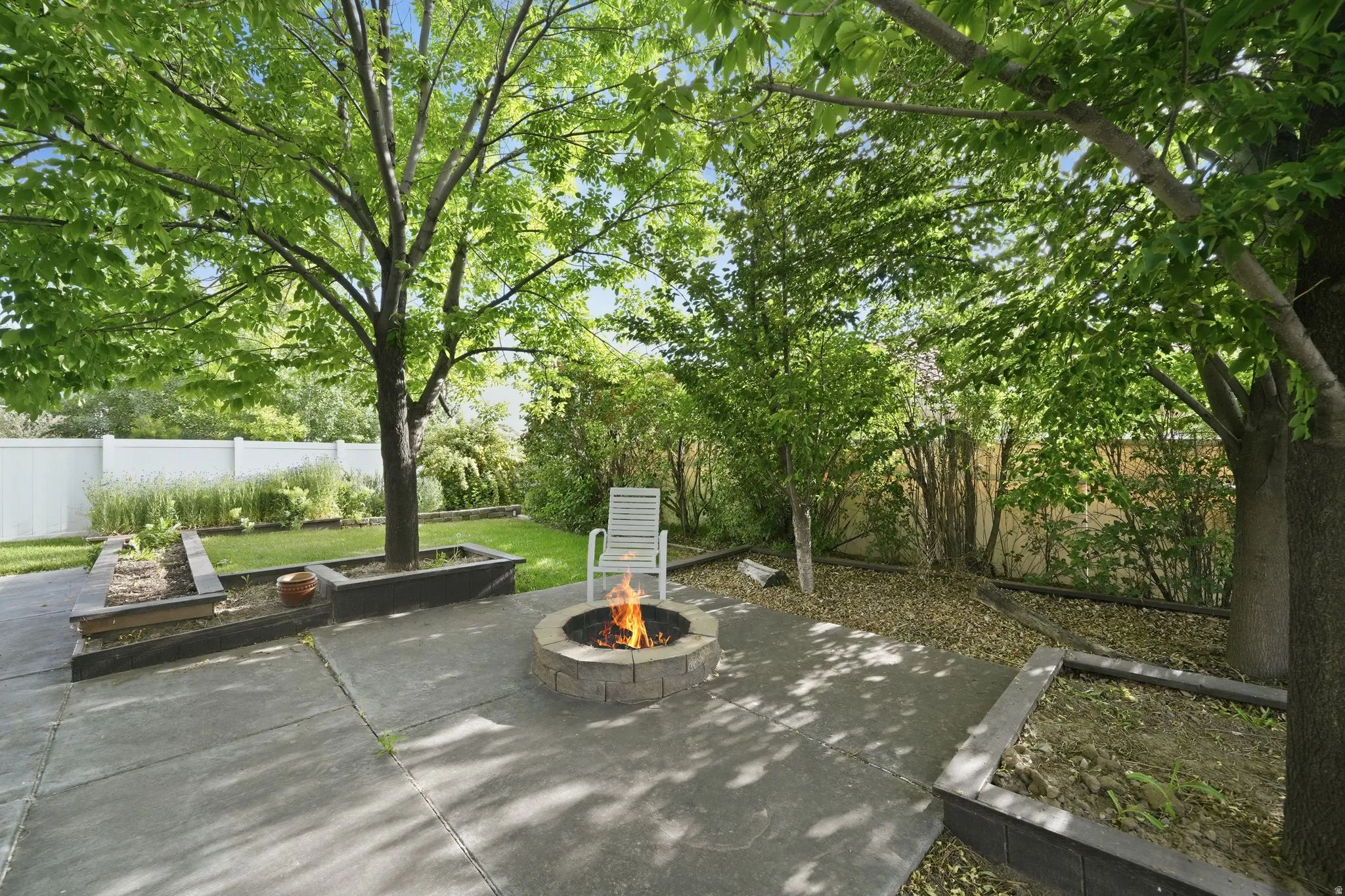 Expansive backyard featuring a concrete patio, a stone-finish fire pit, raised garden beds, mature trees, and a white privacy fence