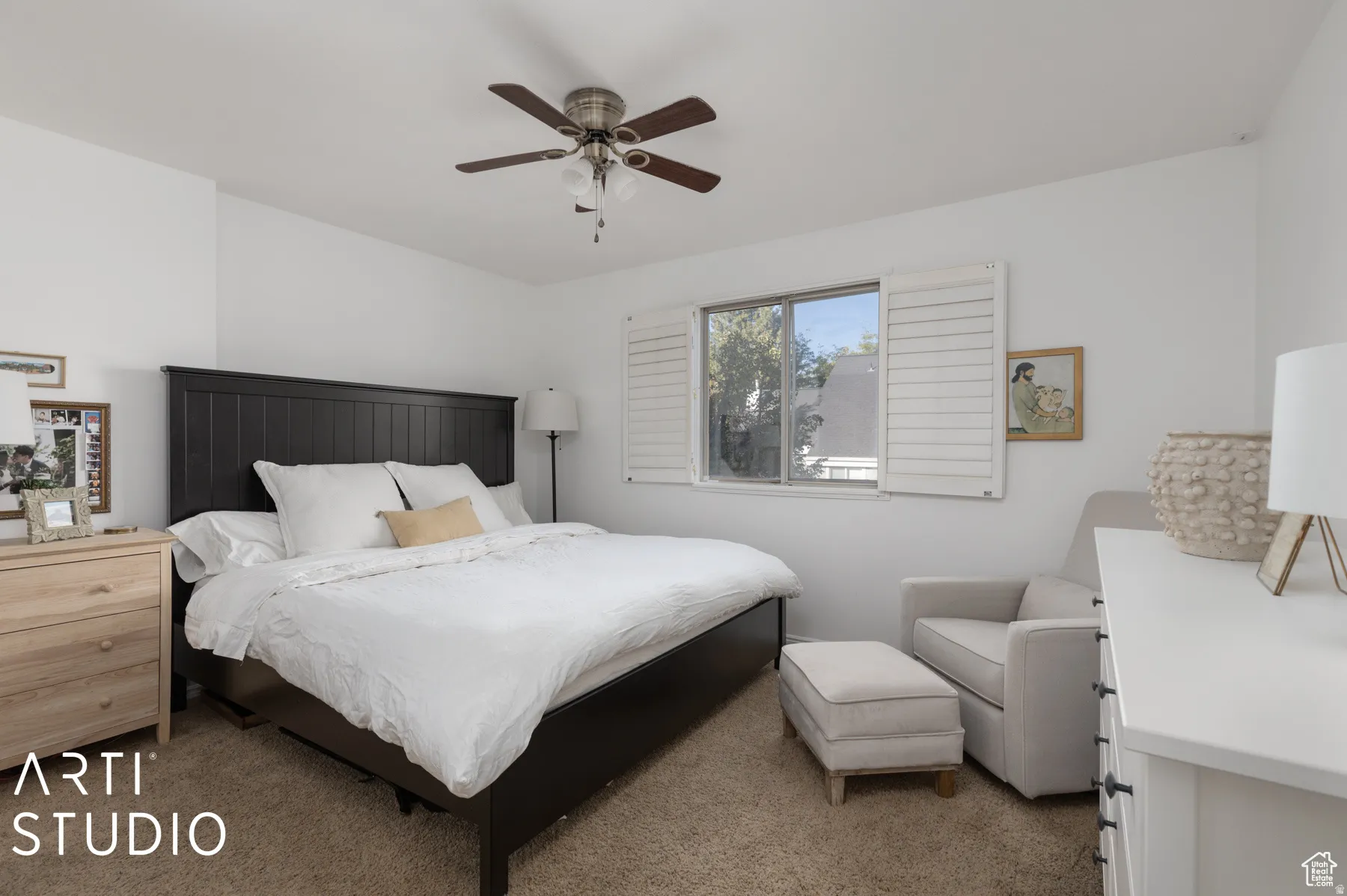 Carpeted bedroom featuring a ceiling fan with light fixture, window with white plantation shutters, and light-colored walls