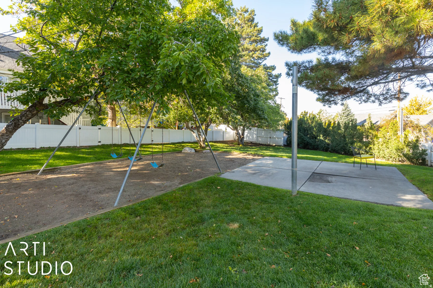Expansive green space featuring a swing set, a paved court area, mature trees, and a white privacy fence