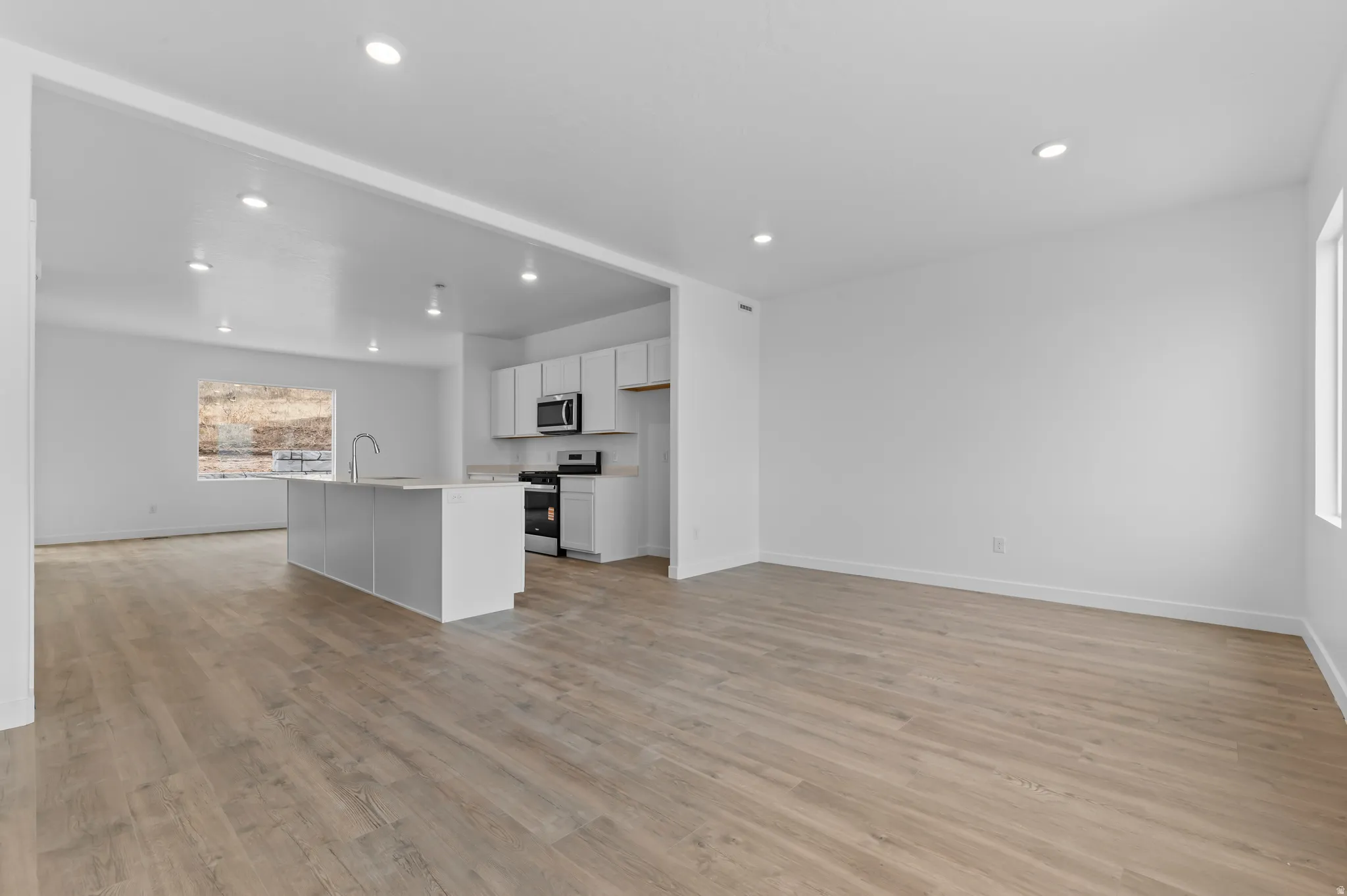 Open-concept living area featuring wood-finish flooring, recessed lighting, and a kitchen with white cabinetry, a central island with a sink, and stainless steel appliances