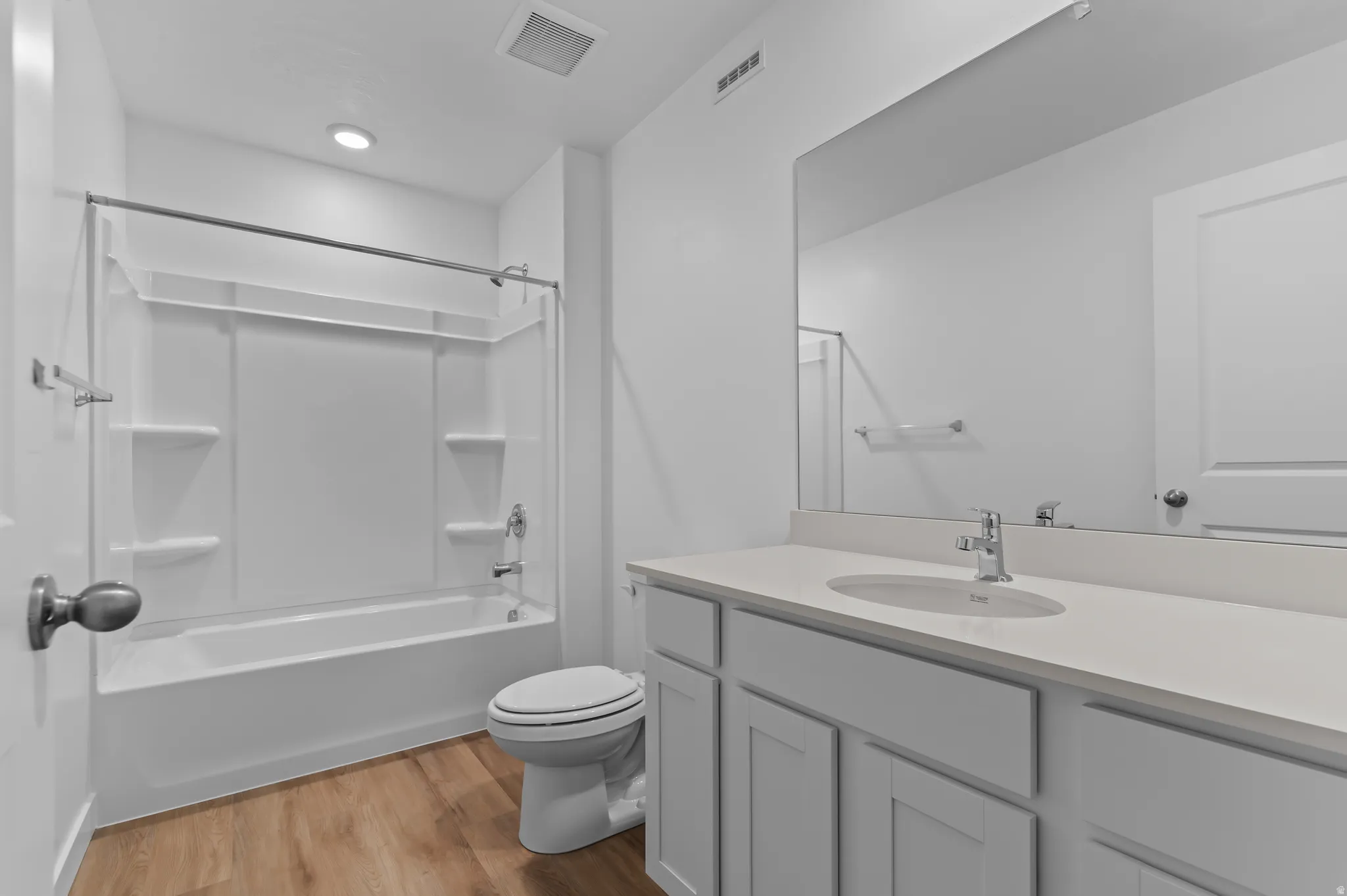 Contemporary bathroom featuring a white vanity with an integrated sink, a large wall-mounted mirror, a tub/shower combination with a white surround, and wood-finish flooring