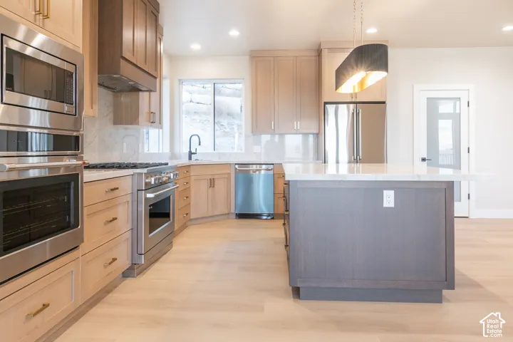 Gourmet kitchen with stainless steel appliances, light wood-finish cabinetry, a central island with a light-toned countertop, and modern pendant lighting