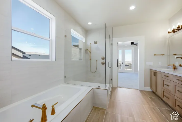 Freestanding soaking tub with gold-tone fixtures, glass-enclosed shower, dual vanity with quartz countertop, light wood-finish cabinetry, and neutral tile flooring