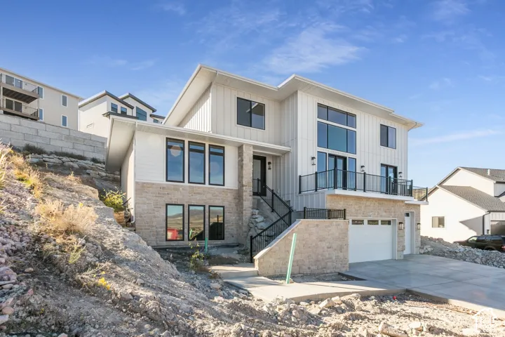 Modern architectural design featuring vertical and horizontal siding, stone facade, multiple windows with black frames, a balcony with a black railing, and an attached garage