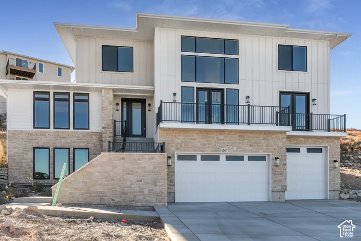 Modern exterior featuring stone and vertical siding accents, multiple balconies with black railings, a prominent main entrance with a dark door, and a two-car garage