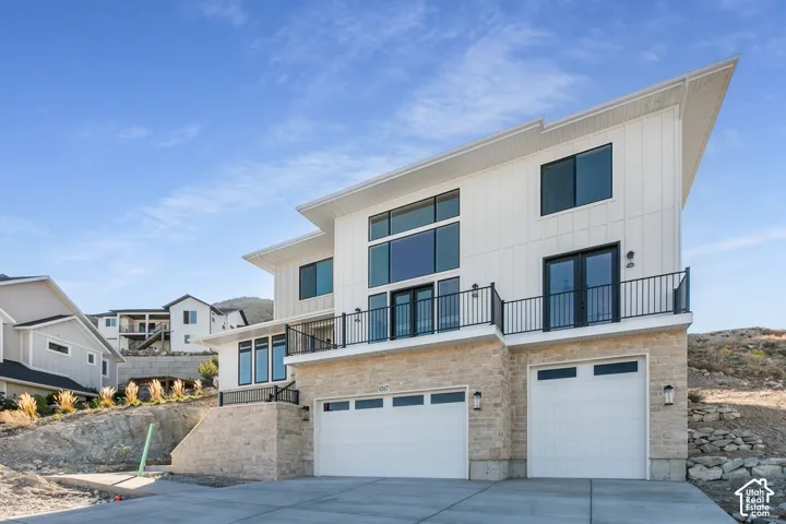 Contemporary exterior with vertical siding, stone veneer accents, multiple large windows, a second-story balcony with metal railings, and two garage doors