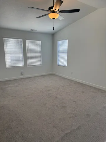 Spacious room featuring vaulted ceilings, a ceiling fan with integrated lighting, three windows with horizontal blinds, and neutral-toned carpeting