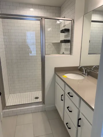 Bathroom featuring a glass-enclosed shower with white subway tile surround, a single vanity with a solid surface countertop, white cabinetry with matte black hardware, and large format floor tiling
