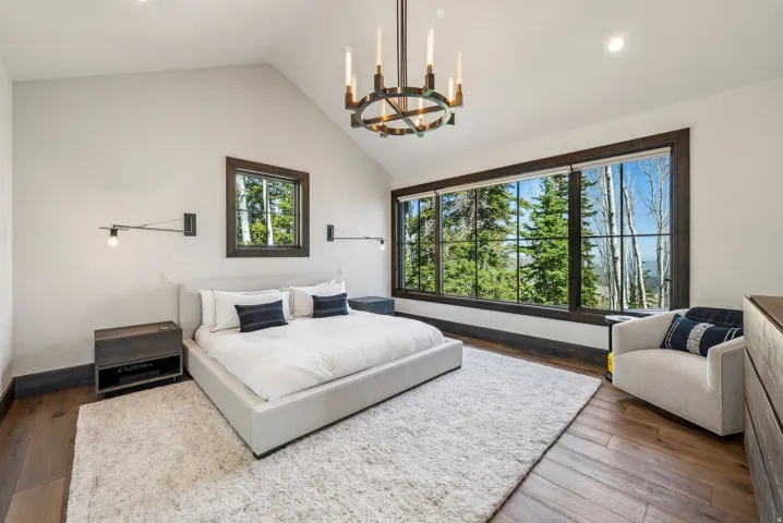 Spacious bedroom featuring vaulted ceilings, a large picture window with dark trim, rich wood-finish flooring, and a contemporary chandelier