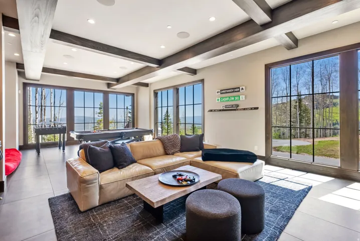 Expansive entertainment room featuring large grid windows, wood-finish ceiling beams, recessed lighting, and tile flooring