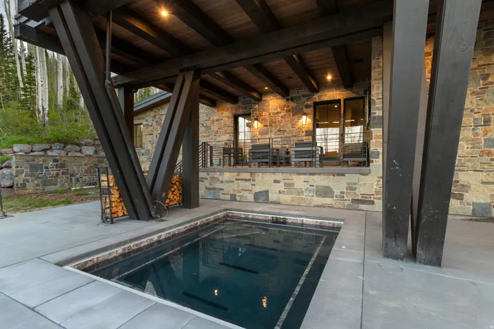 In-ground plunge pool surrounded by a concrete patio, featuring a stone exterior and a covered deck with exposed timber beams and recessed lighting