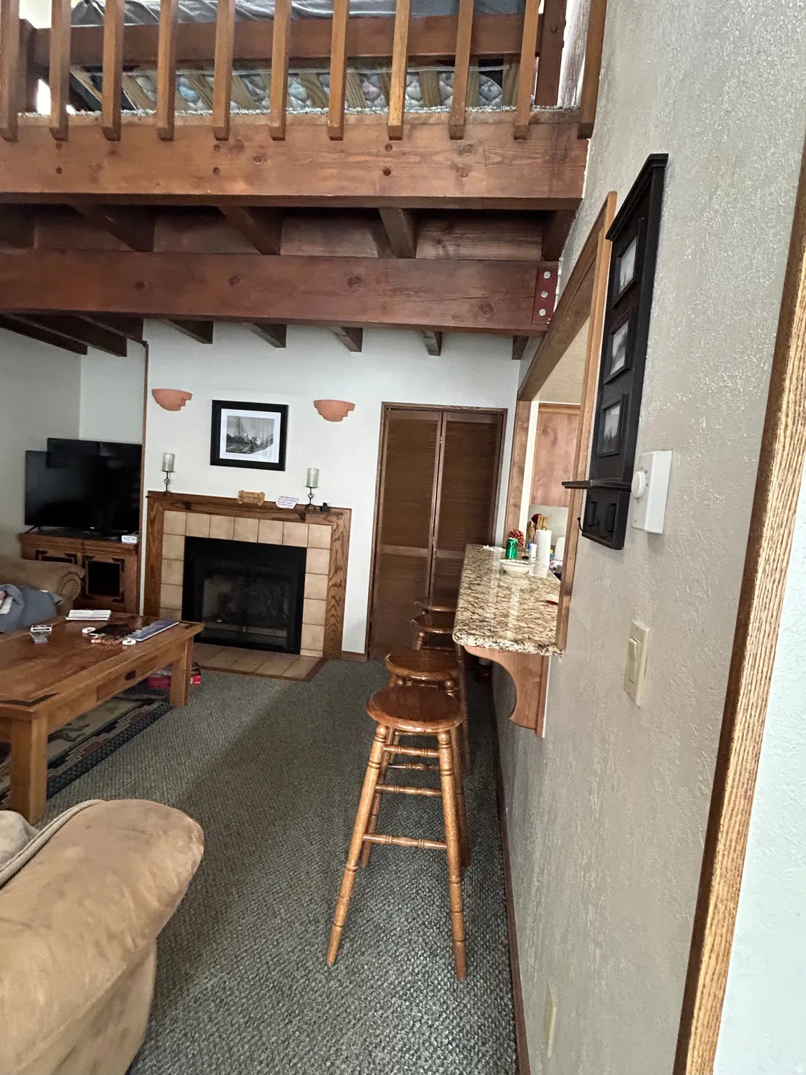 Open-concept living space featuring a wood-burning fireplace with a tile surround and hearth, a breakfast bar with a speckled stone countertop, and exposed timber beams supporting a lofted area with a wood railing