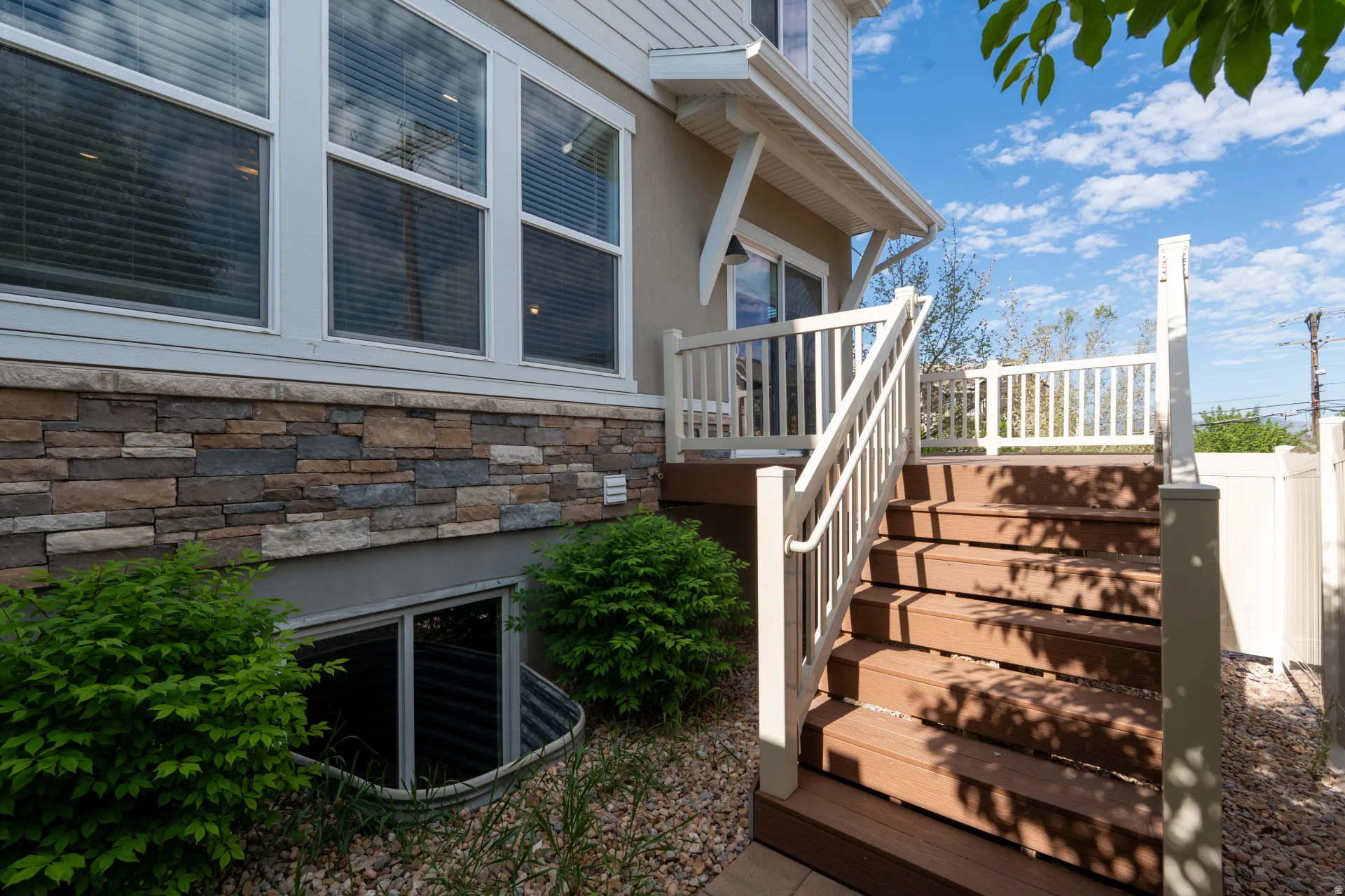 Exterior featuring a composite deck with white railings and stairs, a stone veneer foundation, multiple large windows with blinds, a sliding glass door, and a basement window well