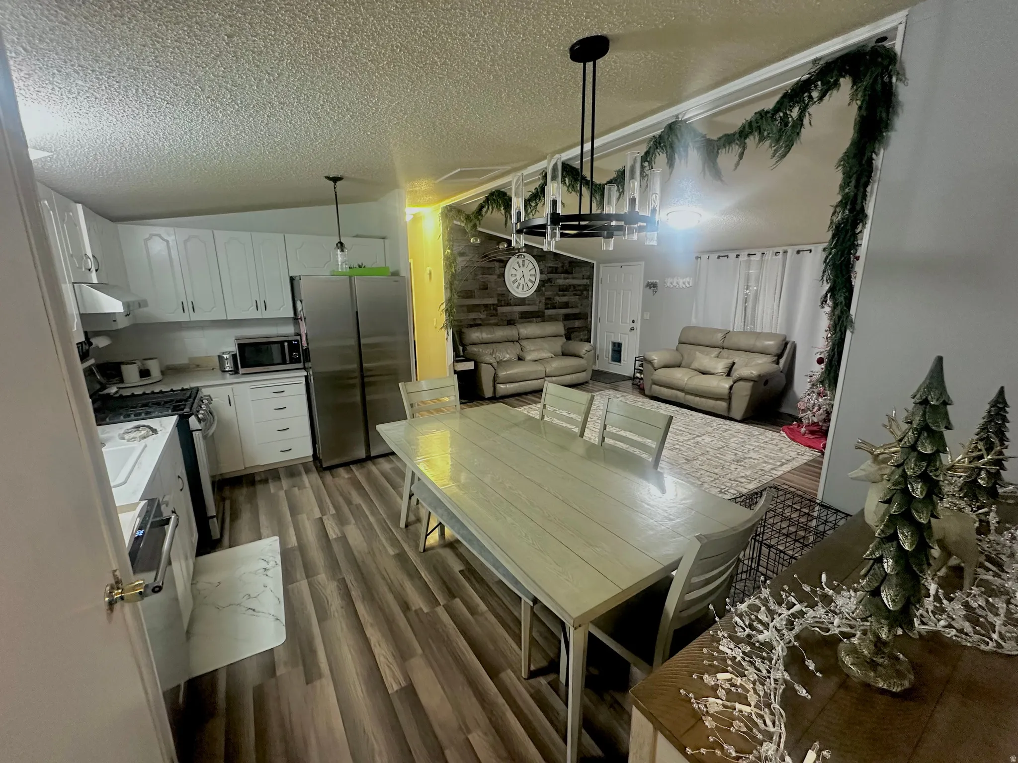 Open-concept living space featuring wood-finish flooring throughout, a dining area with a rectangular table, and an adjacent kitchen with white cabinetry and stainless steel appliances