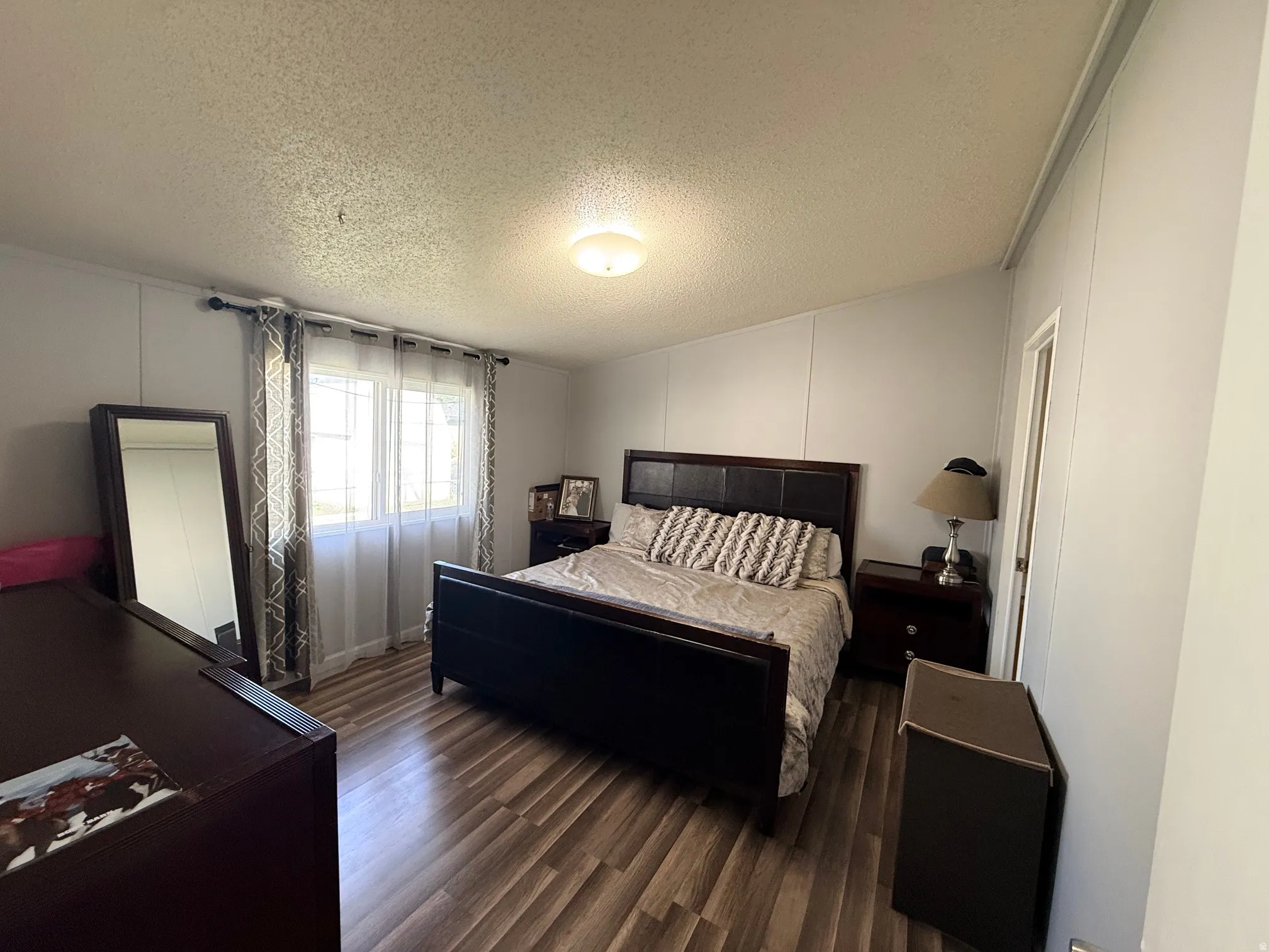 Spacious room featuring wood-finish flooring, light grey paneled walls, and a large window with privacy sheers