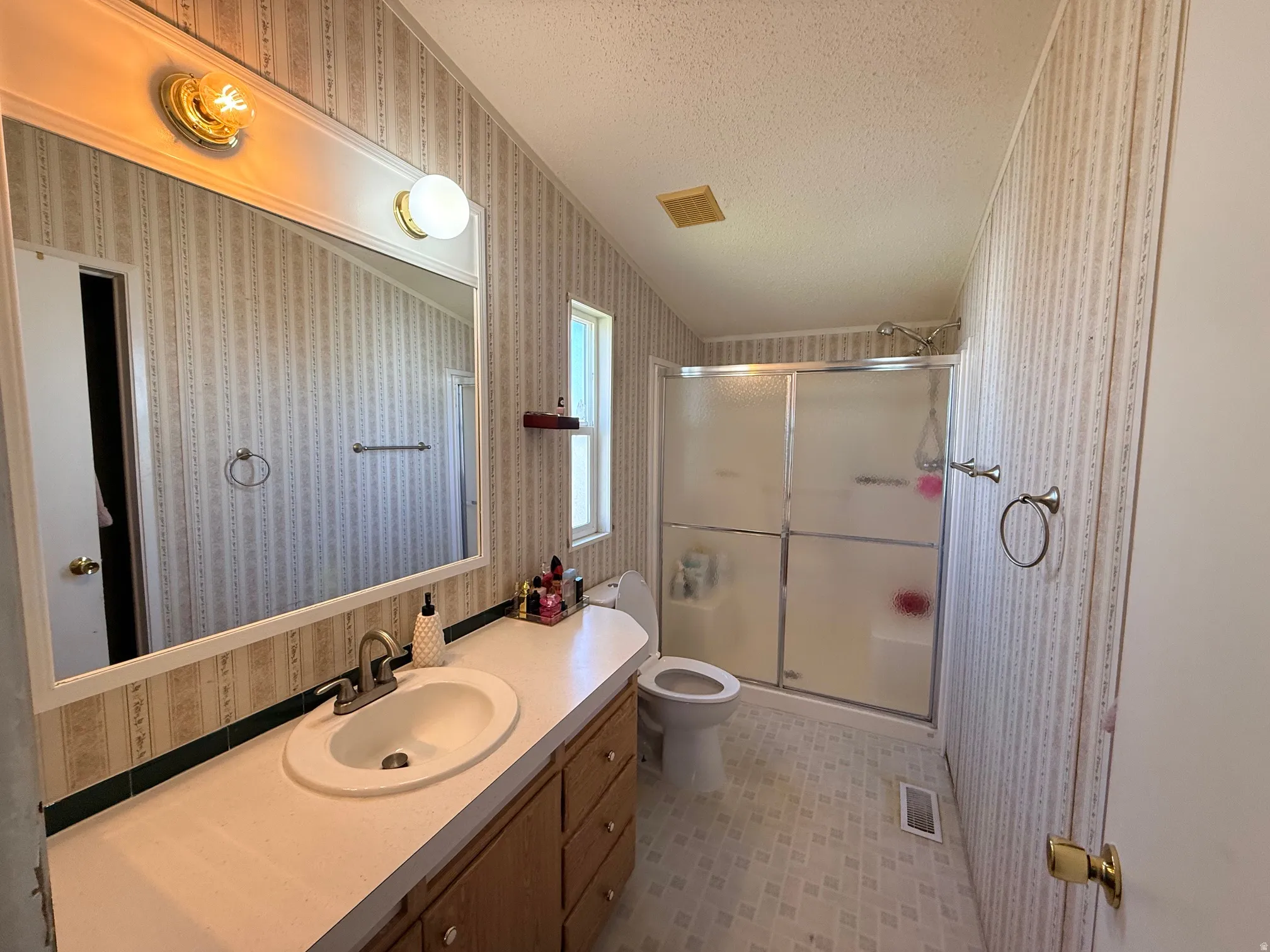 Bathroom featuring a wood-finish vanity with a built-in sink, a large wall-mounted mirror, overhead vanity lighting, a shower enclosure with frosted glass panels, and patterned wall covering