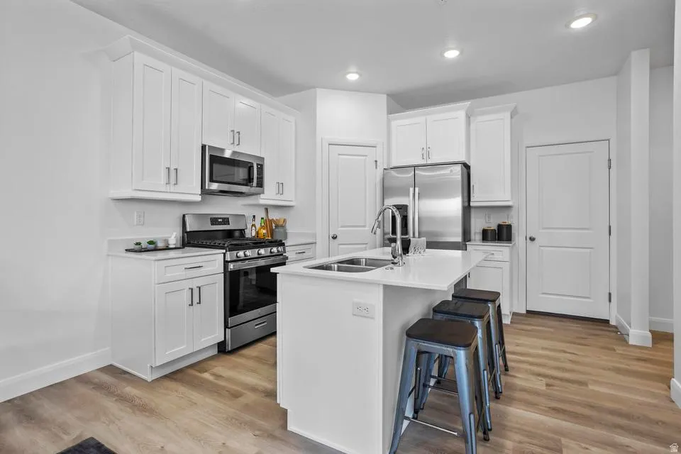 Modern kitchen featuring white cabinetry, stainless steel appliances, a center island with a double sink, light-toned countertops, and wood-finish flooring