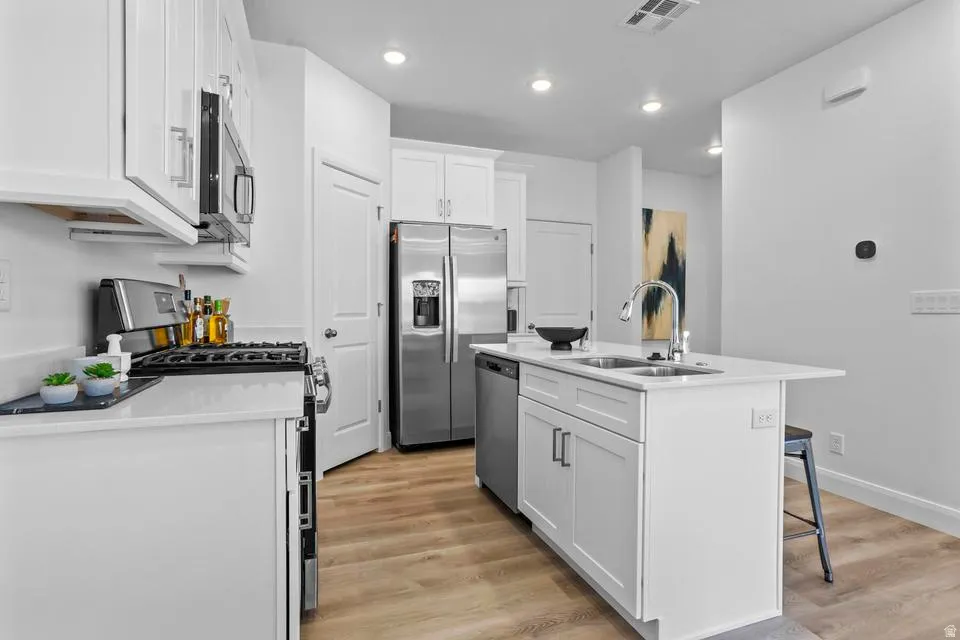 Modern kitchen featuring white shaker cabinetry, stainless steel appliances, light-toned wood-finish flooring, recessed lighting, and a kitchen island with an integrated sink and faucet
