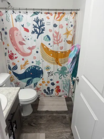 Full bathroom featuring a shower-tub combination with a chrome showerhead, a white toilet, a vanity with a light-toned countertop, dark wood-finish cabinetry with black hardware, and wood-finish flooring