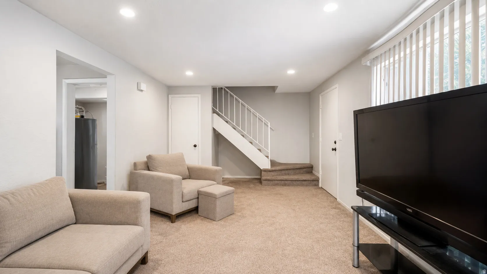 Spacious carpeted room featuring recessed lighting, a staircase with white railings, and a window with vertical blinds