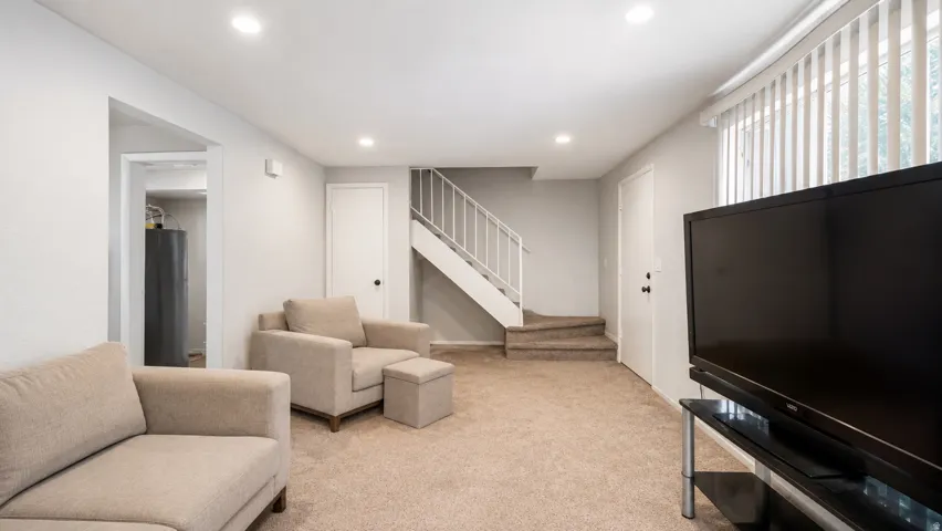 Spacious carpeted room featuring recessed lighting, a staircase with white railings, and a window with vertical blinds