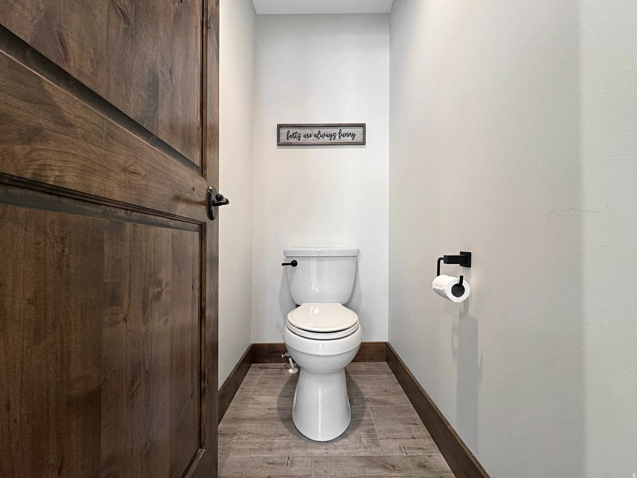 White toilet with a dark-finish flush lever, wood-finish flooring, dark wood baseboards, and a dark wood door with dark hardware